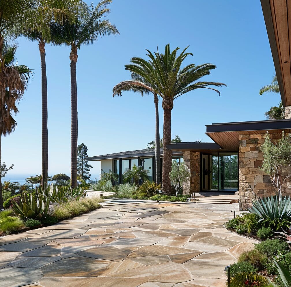 A modern house with stone walls and a large stone patio, surrounded by tropical plants and palm trees, with a view of the ocean in the background.