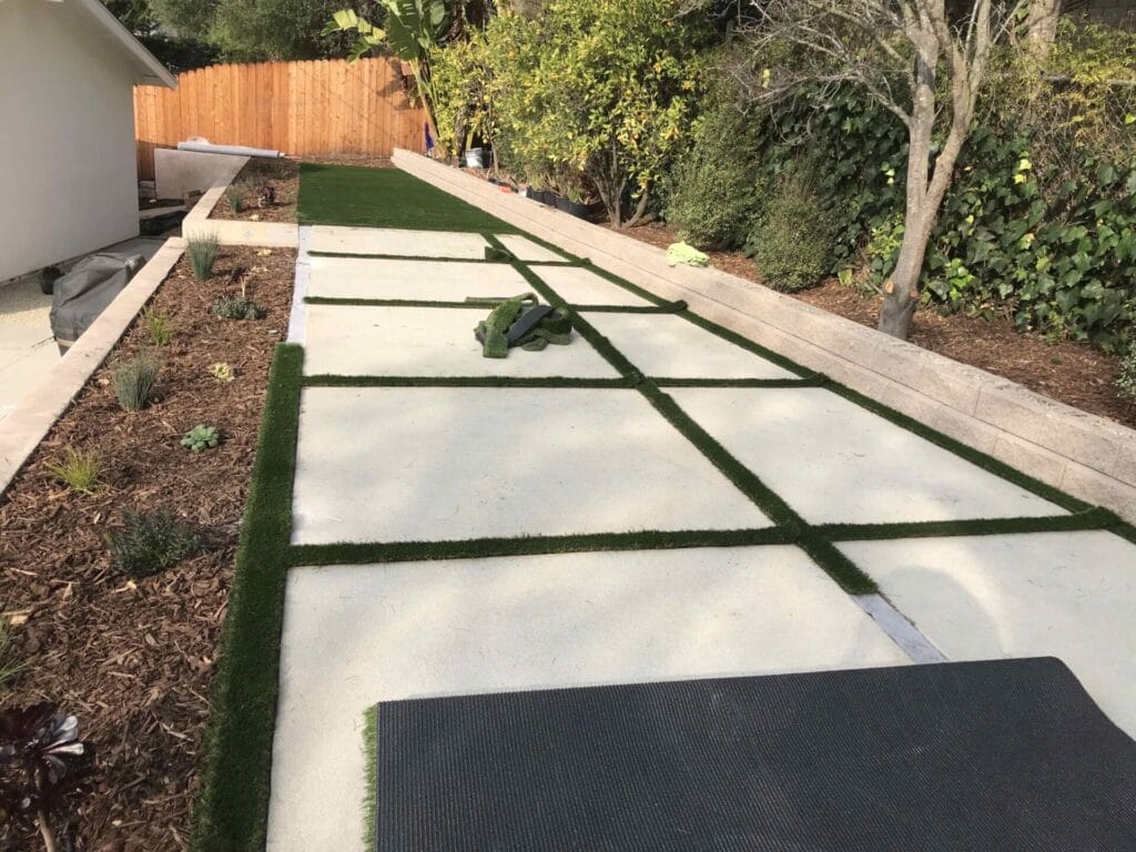A backyard walkway under construction with concrete slabs and artificial grass strips, bordered by mulch and a wooden fence. Tools and materials are visible on the ground.