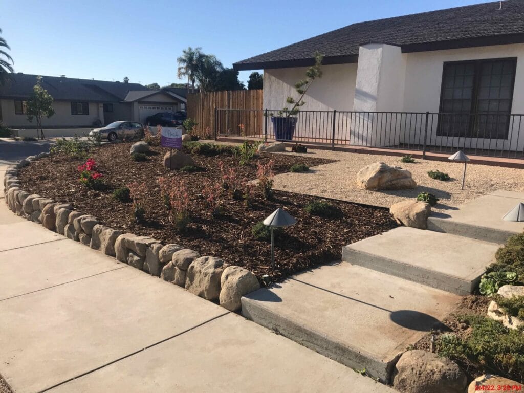 A suburban home with a minimalist xeriscape front yard featuring mulch, scattered plants, rocks, and pathway lighting. Steps lead up to the entrance, and neighboring houses are visible in the background.